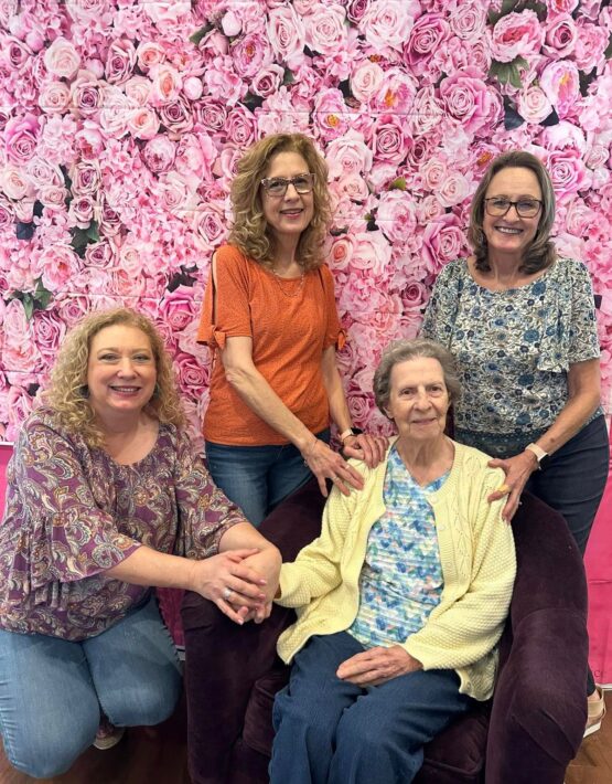 Assisted living senior resident seated with three women standing beside her, all smiling in front of a pink floral backdrop during a community event.