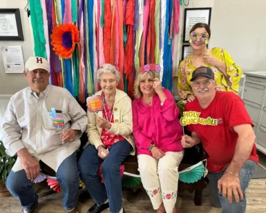 Group of assisted living residents and staff smiling and posing with festive props in front of a colorful decorated backdrop during a community celebration.