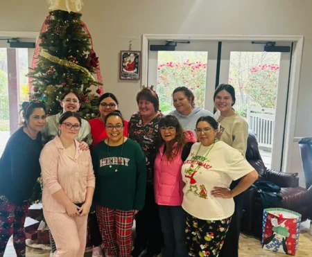 Group of assisted living senior residents and staff smiling together in front of a decorated Christmas tree during a festive holiday gathering.
