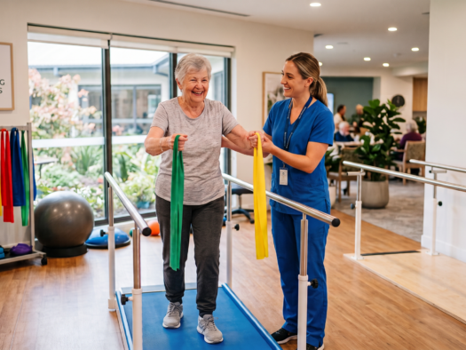 An elderly woman with a walker uses resistance bands under the guidance of a physical therapist.